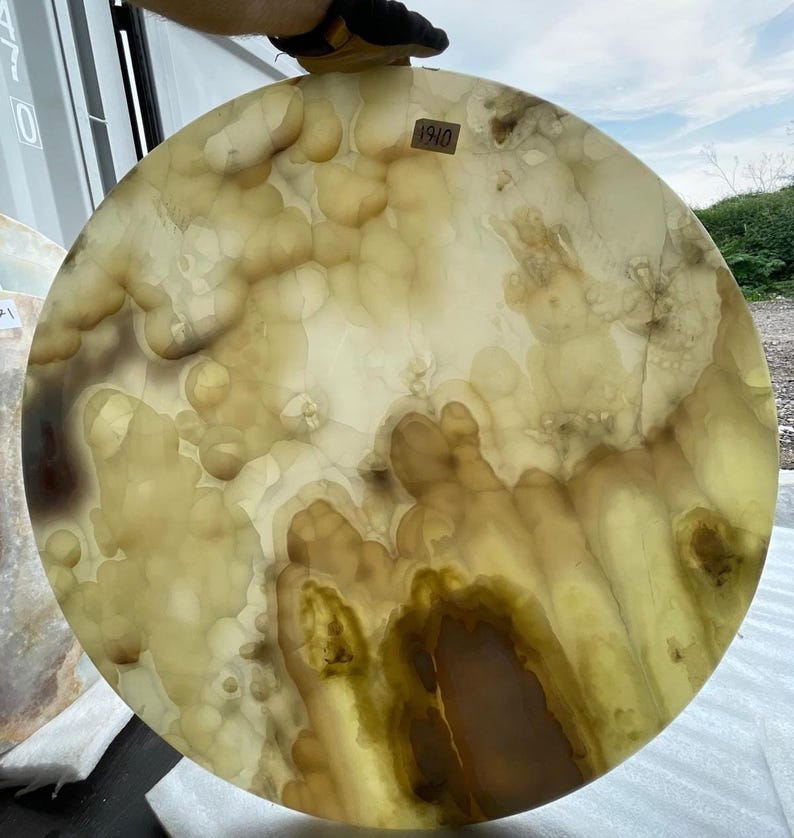 May include: A large, round, translucent stone slab with a unique pattern of light yellow, brown, and white swirls. The stone has a polished surface and a natural, organic appearance. The number "1910" is visible on a small sticker.