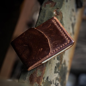Puede incluir: Una cartera de cuero marrón con un aspecto desgastado y texturizado. La cartera tiene un bolsillo curvo en la parte delantera y está cosida a lo largo de los bordes. La cartera descansa sobre una superficie metálica oxidada y desgastada.