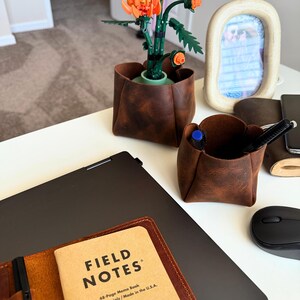 Peut inclure: Sc&egrave;ne de bureau avec un porte-stylo en cuir marron, un ordinateur portable et un carnet "FIELD NOTES". Une composition florale Lego orange et verte est dans un pot en cuir marron. Une souris d'ordinateur noire et un cadre photo sont &eacute;galement visibles.