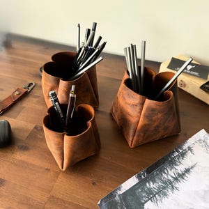 Könnte beinhalten: Drei braune Schreibtisch-Organizer aus Leder in verschiedenen Größen, jeweils mit einem einzigartigen vierblättrigen Design. Die Organizer sind mit Stiften und Bleistiften gefüllt. Das Leder hat ein Used-Finish, was zur rustikalen Ästhetik beiträgt. Ein stilvolles und funktionelles Schreibtischzubehör.