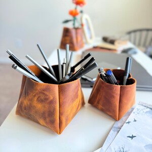 Peut inclure: Deux organisateurs de bureau en cuir marron, un grand et un petit, sont pos&eacute;s sur une surface blanche. Les organisateurs sont remplis de stylos et de crayons. En arri&egrave;re-plan, un vase avec des fleurs oranges est visible.