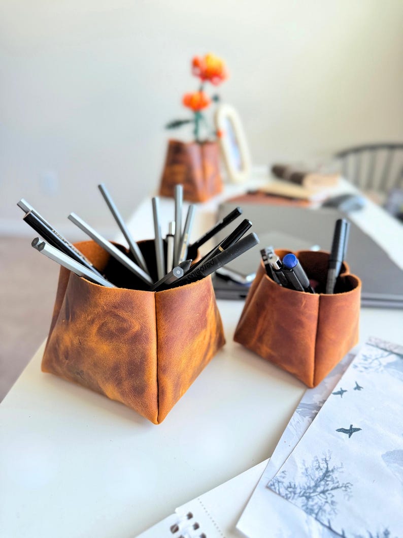 Peut inclure: Deux organisateurs de bureau en cuir marron, un grand et un petit, sont pos&eacute;s sur une surface blanche. Les organisateurs sont remplis de stylos et de crayons. En arri&egrave;re-plan, un vase avec des fleurs oranges est visible.