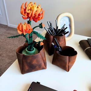 Peut inclure: Une composition de bureau avec une fausse fleur orange et jaune dans un pot en cuir marron, ainsi que deux autres r&eacute;cipients en cuir contenant des stylos et des crayons. Un ordinateur portable et une photo encadr&eacute;e sont &eacute;galement visibles.