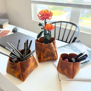 Peut inclure: Trois organiseurs de bureau en cuir marron de diff&eacute;rentes tailles sont pos&eacute;s sur un bureau blanc. Ils contiennent des crayons, des stylos et une composition florale orange. Le bureau est pr&egrave;s d'une fen&ecirc;tre avec des stores.