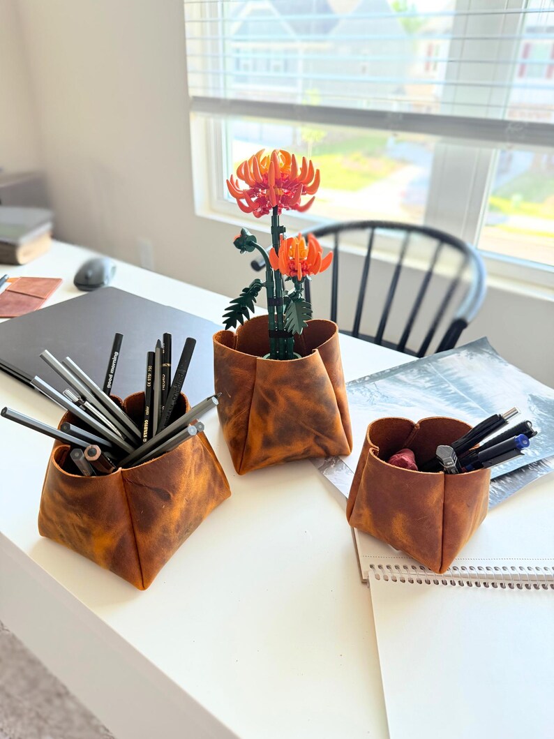 Peut inclure: Trois organiseurs de bureau en cuir marron de diff&eacute;rentes tailles sont pos&eacute;s sur un bureau blanc. Ils contiennent des crayons, des stylos et une composition florale orange. Le bureau est pr&egrave;s d'une fen&ecirc;tre avec des stores.