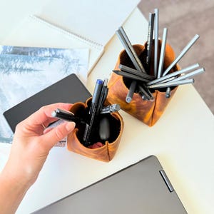 Peut inclure: Deux organiseurs de bureau en cuir marron remplis de stylos et de crayons. Un organisateur est tenu par une personne. Un ordinateur portable, un carnet et d'autres articles de papeterie sont sur le bureau blanc.