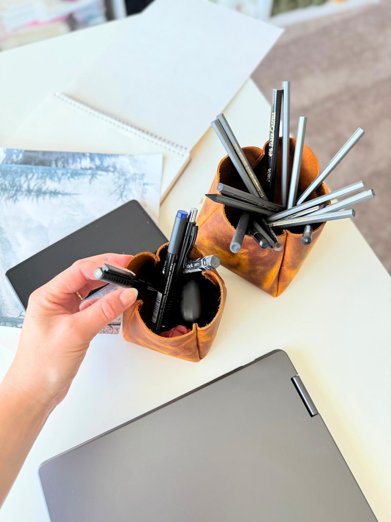 Peut inclure: Deux organiseurs de bureau en cuir marron remplis de stylos et de crayons. Un organisateur est tenu par une personne. Un ordinateur portable, un carnet et d'autres articles de papeterie sont sur le bureau blanc.