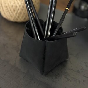 Peut inclure: Un organiseur de bureau en cuir noir contenant plusieurs crayons noirs. L'organisateur a une forme g&eacute;om&eacute;trique unique. Une pelote de ficelle est visible en arri&egrave;re-plan.