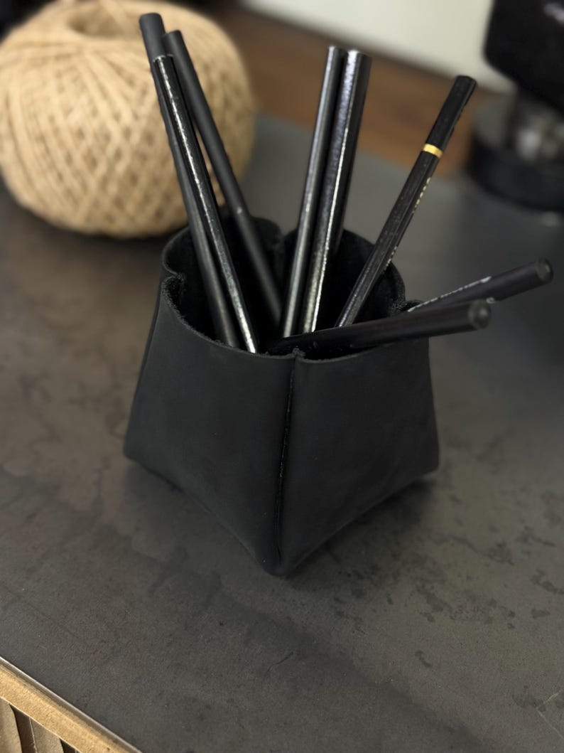Peut inclure: Un organiseur de bureau en cuir noir contenant plusieurs crayons noirs. L'organisateur a une forme g&eacute;om&eacute;trique unique. Une pelote de ficelle est visible en arri&egrave;re-plan.