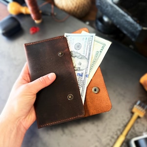 Puede incluir: Una cartera de cuero marrón oscuro con cierre de botón a presión, que muestra varios billetes de dólares estadounidenses. La cartera es de un tono marrón oscuro y parece hecha a mano. La cartera está en la mano de una persona.