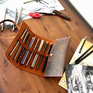 May include: A brown leather pencil case, open to display various pencils, rests on a wooden surface. The case is surrounded by art supplies, including a sketchbook, eraser, and a small knife. The pencils are of different colors and sizes.