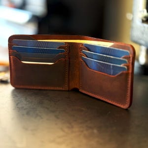 May include: Open brown leather wallet with multiple card slots filled with blue and white striped cards. The wallet is open, showcasing the interior design and the cards within. The leather has a textured appearance.