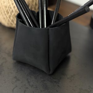 Peut inclure: Un porte-crayons en cuir noir rempli de plusieurs crayons noirs. Le support est de forme carr&eacute;e avec un design pli&eacute;. Une pelote de ficelle est visible en arri&egrave;re-plan, sugg&eacute;rant un cadre artisanal ou de bureau.