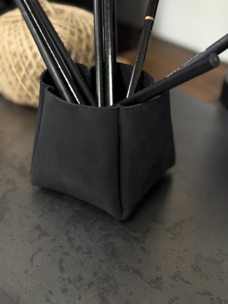Peut inclure: Un porte-crayons en cuir noir rempli de plusieurs crayons noirs. Le support est de forme carr&eacute;e avec un design pli&eacute;. Une pelote de ficelle est visible en arri&egrave;re-plan, sugg&eacute;rant un cadre artisanal ou de bureau.