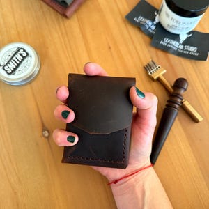 May include: A dark brown leather wallet with a curved flap, held in a hand. The wallet is stitched along the edges. Other leather goods and leather care products are in the background.