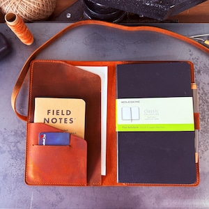 Puede incluir: Funda de cuaderno de cuero marrón abierta, con un bolsillo que contiene un cuaderno Field Notes y una tarjeta. Se ve un cuaderno Moleskine negro con una banda verde. La funda tiene una correa y está sobre una superficie metálica.