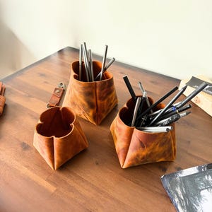 Peut inclure: Trois organiseurs de bureau en cuir marron avec un design unique en forme de p&eacute;tale. Les organiseurs sont remplis de stylos, de crayons et de pailles m&eacute;talliques. Le cuir a une finition vieillie, ce qui leur donne un aspect rustique. Ils sont pos&eacute;s sur une surface en bois.