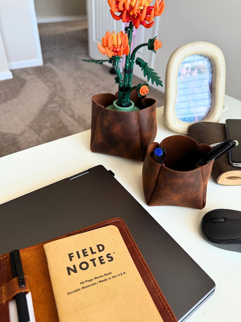 Peut inclure: Sc&egrave;ne de bureau avec un porte-stylo en cuir marron, un ordinateur portable et un carnet "FIELD NOTES". Une composition florale Lego orange et verte est dans un pot en cuir marron. Une souris d'ordinateur noire et un cadre photo sont &eacute;galement visibles.