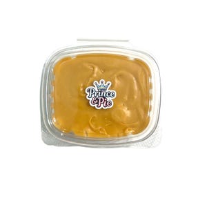 May include: A clear plastic square container filled with a smooth, light brown caramel-colored substance. A white sticker is centered on the lid featuring a blue crown icon above the text Prince & Pie in blue and pink stylized lettering.