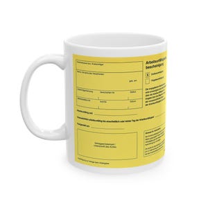 May include: A white ceramic mug featuring a yellow rectangular graphic printed on its side. The graphic replicates a German medical certificate, known as an Arbeitsunfähigkeitsbescheinigung, with black text, lines, and boxes for data entry.
