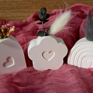 Könnte beinhalten: Drei kleine, weiße dekorative Vasen in Herz-, Blumen- und Regenbogenform. Jede Vase hat einen Herz-Ausschnitt und hält Kunstblumen oder dekorative Stiele. Die Vasen sind auf einem strukturierten, rosa Stoff angeordnet.