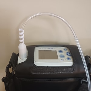 May include: A portable oxygen concentrator with a black carrying case and a clear oxygen tube. The device has a digital display with the text "INOGEN ONE" and buttons for control.