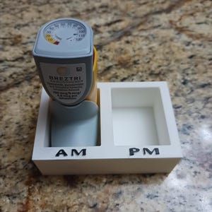 May include: A Breztri Aerosphere inhaler with a built-in dose counter, resting in a white holder labeled "AM" and "PM". The inhaler is gray, yellow, and blue, with text indicating the medication and dosage.