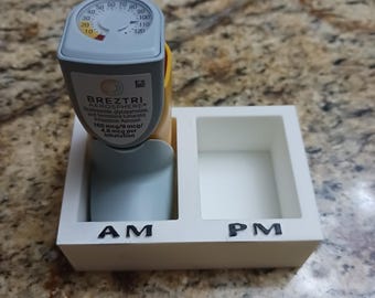 May include: A Breztri Aerosphere inhaler with a built-in dose counter, resting in a white holder labeled "AM" and "PM". The inhaler is gray, yellow, and blue, with text indicating the medication and dosage.