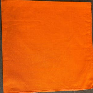 May include: Bright orange square fabric, likely a bandana or handkerchief, with a textured surface. The fabric is folded neatly, showcasing its vibrant color and simple design. The edges are finished with a stitched hem.