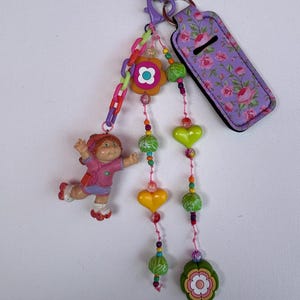 Peut inclure: Un porte-clés fantaisiste avec une petite poupée, des perles colorées et une étiquette à motifs floraux. Le porte-clés comprend une chaîne, un charm en forme de fleur et des perles en forme de cœur. L'étiquette est violette avec un motif de roses roses.