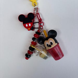May include: A Mickey Mouse-themed keyring featuring a yellow loop, a red and black Mickey Mouse head, and a red cup with a Mickey Mouse face. Also includes a clear boba drink cup and a beaded charm.
