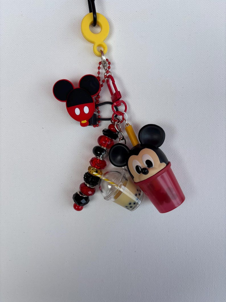 May include: A Mickey Mouse-themed keyring featuring a yellow loop, a red and black Mickey Mouse head, and a red cup with a Mickey Mouse face. Also includes a clear boba drink cup and a beaded charm.
