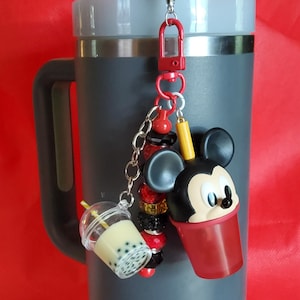 May include: A grey tumbler with a Mickey Mouse-themed charm. The charm features a Mickey Mouse head, a boba tea cup, and decorative beads. The tumbler has a black handle and a red background.