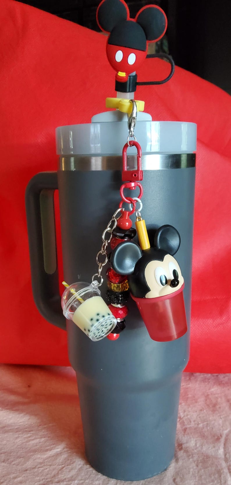 May include: A grey tumbler with a Mickey Mouse-themed charm. The charm features a Mickey Mouse head, a boba tea cup, and decorative beads. The tumbler has a black handle and a red background.
