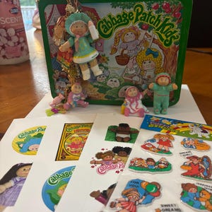 Peut inclure: Une boîte à lunch en métal vert Cabbage Patch Kids avec une illustration colorée de personnages, ornée d'un bracelet à breloques avec de petites figurines. En dessous se trouvent plusieurs feuilles d'autocollants et décalcomanies Cabbage Patch Kids.