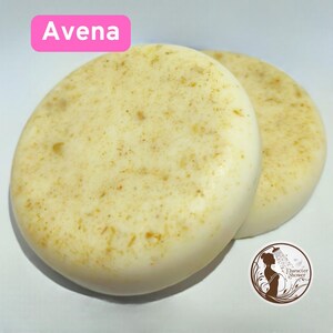 Puede incluir: Dos jabones redondos y claros con una textura moteada. Los jabones tienen un aspecto cremoso con motas marrones. La palabra "Avena" se muestra en un rectángulo rosa en la parte superior. Un logotipo con una mujer bajo la ducha está en la esquina inferior derecha.