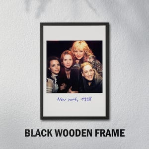 May include: A framed photograph featuring four women, with the handwritten text "New york, 1998" in blue ink. The photograph is set within a black wooden frame, mounted on a white wall.
