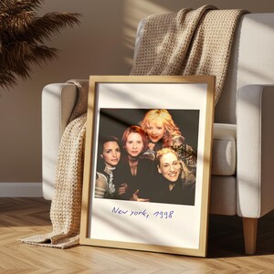 May include: A framed photograph featuring four women, with the handwritten text "New york, 1998" at the bottom. The frame is light-colored wood, and the photograph is propped against a chair with a beige throw blanket.