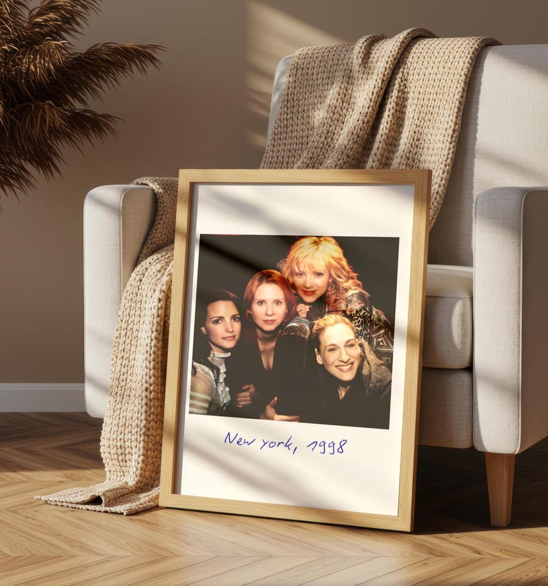 May include: A framed photograph featuring four women, with the handwritten text "New york, 1998" at the bottom. The frame is light-colored wood, and the photograph is propped against a chair with a beige throw blanket.