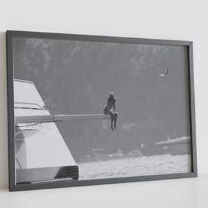 Op de afbeelding: Zwart-wit ingelijste print van een persoon die op de rand van een boot zit, met een vliegende zeemeeuw op de achtergrond. De boot bevindt zich op het water en de afbeelding heeft een serene, minimalistische uitstraling.