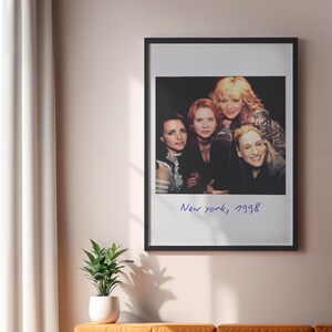 May include: A framed print featuring a group of four women. The image is in a black frame and has the text "New york, 1998" in a handwritten style. The print is hanging on a light pink wall.
