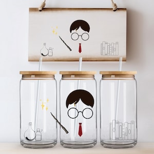Op de afbeelding: Drie doorzichtige glazen bekers met bamboe deksels en rietjes, elk met een andere zwarte afbeelding. Eén heeft een drankfles, een andere een toverstaf en sterren, en de laatste een stapel boeken. Een bijpassend bord hangt erboven.