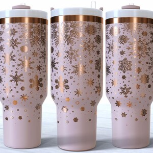 May include: Three insulated tumblers with a light pink base and a white lid. The tumblers feature a gold snowflake pattern and a matching handle. The rim is accented with a gold band. The tumblers are approximately 25 cm tall.