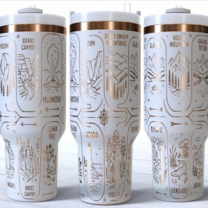 Puede incluir: Taza de viaje blanca con asa y tapa, decorada con ilustraciones doradas de monumentos de parques nacionales. La taza presenta nombres de parques como Yosemite, Grand Canyon y Yellowstone. El diseño envuelve la taza.