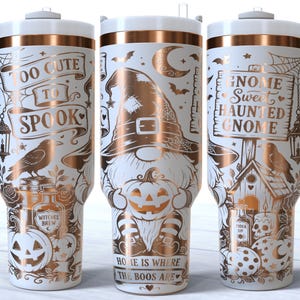 Könnte beinhalten: Drei weiße Becher mit Henkeln und kupferfarbenen Halloween-Motiven. Die Designs zeigen einen Zwerg mit Hexenhut, der einen Kürbis hält, einen Raben, ein Spinnennetz sowie den Text "TOO CUTE TO SPOOK," "GNOME SWEET HAUNTED GNOME," und "HOME IS WHERE THE BOOS ARE."