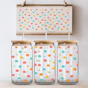 May include: Three clear glass drinking jars with bamboo lids and straws, decorated with colorful floral patterns. A matching floral design is displayed on a wooden sign in the background. The flowers are in shades of red, yellow, orange, and blue.