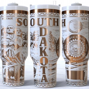May include: White travel mug with gold accents and a handle. The mug features illustrations of Mount Rushmore, a bison, and the words "South Dakota" and "USA" in gold lettering. The mug has a white lid and handle.
