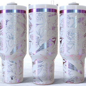 May include: White tumbler with a wraparound design featuring birds and floral elements in a delicate, iridescent finish. The tumbler has a white handle and lid with a metallic band. The design includes roses, lilies, and other flowers.