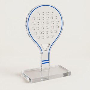 May include: A clear acrylic padel racket display stand featuring a circular head with a grid of perforated holes, a triangular cutout above the handle, and blue line accents along the perimeter and handle base, mounted on a rectangular base.