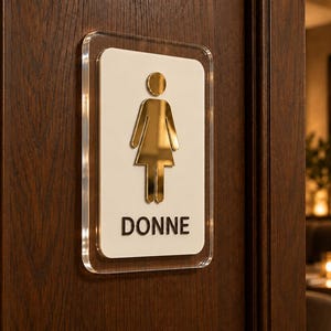 May include: A rectangular restroom sign with a clear acrylic backing, a white center panel, and a gold-toned metallic female figure icon. The word DONNE is printed in black capital letters at the bottom of the white panel, mounted on a dark wood door.